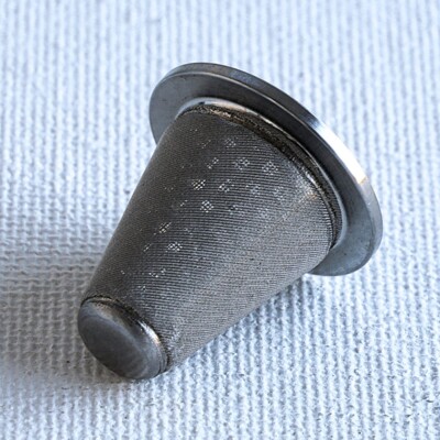 A metal mesh cone tea infuser resting on a textured white surface.